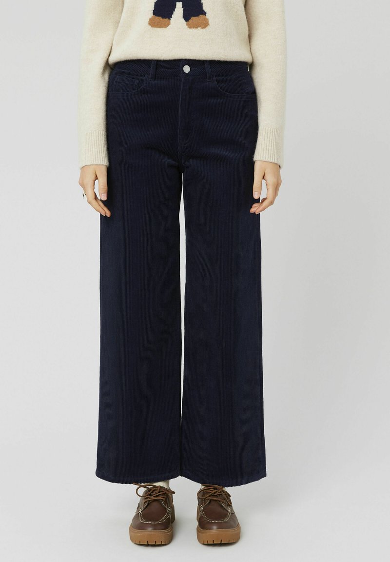 Pantalons larges en velours côtelé bleu marine avec poches avant visibles, fermeture à bouton et nervures texturées ; associés à des chaussures à lacets marron.