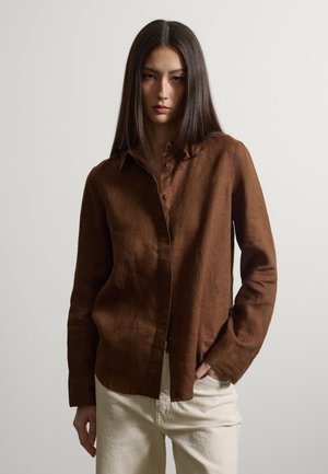 Femme aux cheveux raides et foncés, portant une chemise marron à manches longues boutonnée et un pantalon couleur crème, debout avec une main dans la poche.