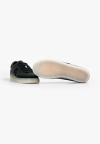 Un par de zapatillas negras de corte bajo con suelas de goma semi-translúcidas, cordones negros y un patrón texturizado en la suela visible en un zapato.