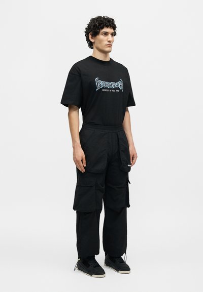 Νέος άνδρας με κατσαρά μαλλιά, φοράει μαύρο μπλουζάκι με γραφικά, μαύρο παντελόνι καρό με πολλές τσέπες και μαύρα αθλητικά παπούτσια, στέκεται σε λευκό φόντο.