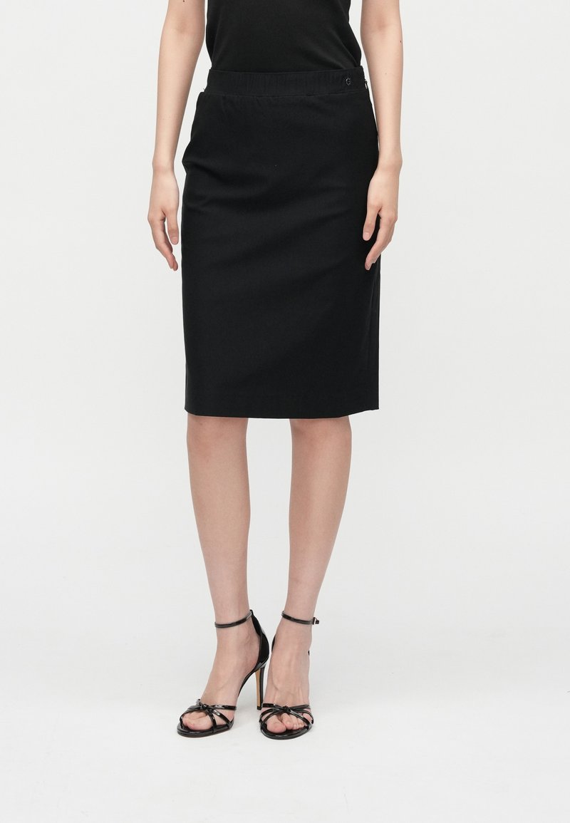 Μαύρη pencil φούστα μέχρι το γόνατο με ελαστική μέση. Λείο ύφασμα, απλός σχεδιασμός, χωρίς ορατά σχέδια ή διακοσμητικά στοιχεία.