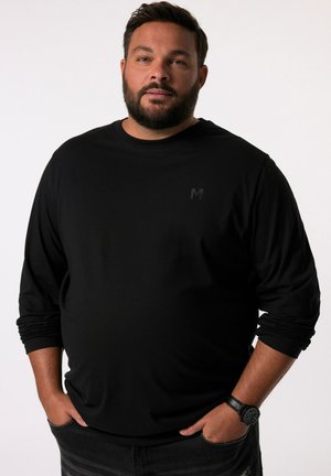 Schwarzes Langarmshirt aus weichem Stoff mit dezentem Logo auf der Brust. Getragen mit dunklen Jeans und einer schwarzen Armbanduhr.