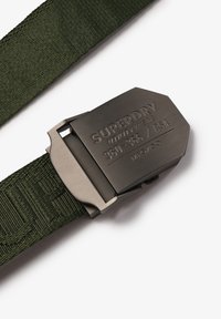Groene gevlochten riem met een metalen gesp waarop "SUPERDRY" en modelgegevens zijn gegraveerd. De gesp heeft een geborstelde afwerking met rechte randen.