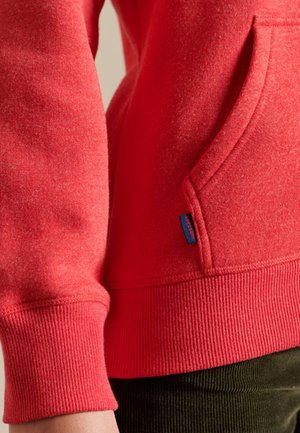 Primer plano de una persona que lleva un sudadera roja con un bolsillo delantero y puños acanalados, combinada con pantalones verde oscuro.