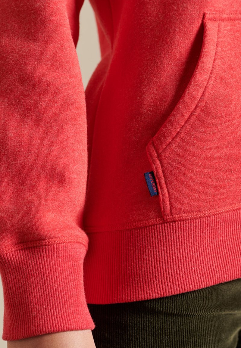 Primer plano de una persona que lleva un sudadera roja con un bolsillo delantero y puños acanalados, combinada con pantalones verde oscuro.