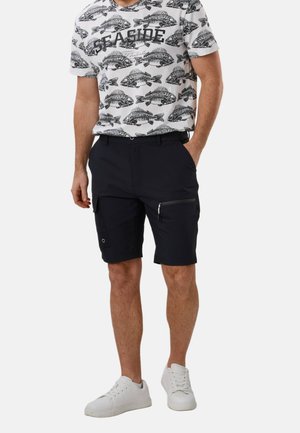 Man wearing a white t-shirt with black fish skeleton print and "SEASIDE" text, black cargo shorts, and white sneakers, standing with one hand in pocket.