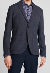 Man som bär en mörk strukturerad blazer över en blå blommig skjorta med knappar och mörka byxor, stående med armarna avslappnat vid sidorna.