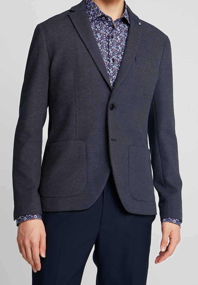 Man som bär en mörk strukturerad blazer över en blå blommig skjorta med knappar och mörka byxor, stående med armarna avslappnat vid sidorna.