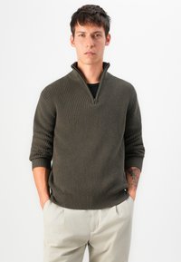 Maglione zipper verde oliva con texture a coste, caratterizzato da un colletto alto e maniche aderenti, abbinato a pantaloni chiari.
