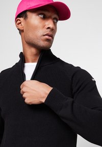 Czarny sweter dziany z kołnierzem na zamek i prążkowaną fakturą, noszony na białej koszuli. Na głowie różowa czapka, która podkreśla stylizację.