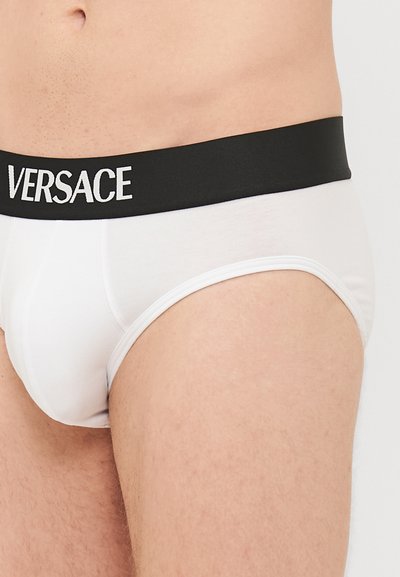 Fehér alsónadrág fekete elasztikus derékpánttal, amelyen fehér betűkkel szerepel a "VERSACE" felirat. Sima textúra, testre simuló fazon és közepes magasító.