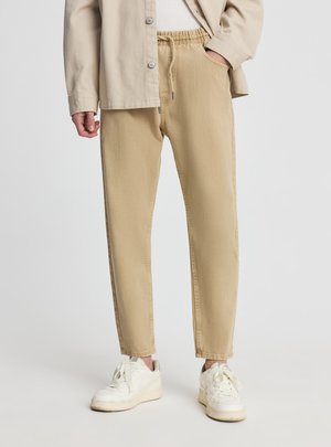 Persona che indossa pantaloni beige con coulisse, camicia crema con bottoni e scarpe da ginnastica bianche, in piedi con una mano in tasca.