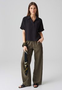 Schwarzes, geripptes Kurzarmhemd mit Kragen, kombiniert mit olivgrünen, weit geschnittenen Hosen und einem gestreiften Schal. Flache Sandalen vervollständigen den Look.