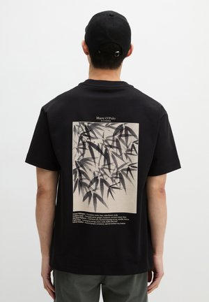 Mężczyzna w czarnym T-shircie z monochromatycznym nadrukiem liścia bambusa i małym blokiem tekstu na plecach, w połączeniu z ciemnymi spodniami i czarną czapką.