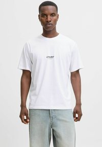 Camiseta de algodón blanca con cuello redondo y mangas cortas, con el logo negro "JJ Studios" bordado en el pecho.
