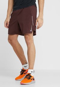 Hombre vestido con pantalones cortos de deporte color burdeos, calcetines blancos de Nike y zapatillas de correr brillantes en color naranja de Nike, de pie contra un fondo liso.