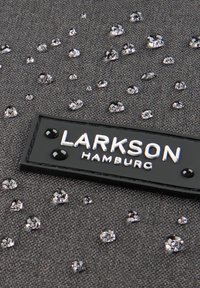 Tela gris con superficie texturizada, adornada con brillantes lentejuelas plateadas y una etiqueta rectangular negra que dice "LARKSON HAMBURG."