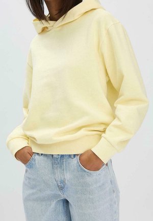 Person trägt einen hellgelben Kapuzenpullover und hellblaue Jeans mit den Händen in den Taschen vor einem einfarbigen Hintergrund.