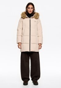 Chaqueta de plumas en beige claro con capucha forrada de piel, cierre de cremallera al frente y dos bolsillos laterales. Corte oversized con textura acolchada.