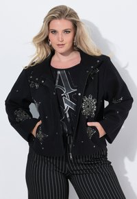 Femme blonde portant une veste noire avec des motifs de perles argentées, un haut graphique noir et un pantalon noir à rayures, debout les mains dans les poches.