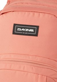 Zaino rosa morbido con un tessuto testurizzato. Presenta un'etichetta nera con "DAKINE" e "EST. 1979." Sono visibili due tasche con zip.