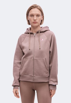 Vrouw met kort blond haar die een mauve zip-up hoodie met voorzakken en bijpassende leggings draagt, staand voor een effen achtergrond.