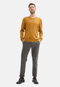 Suéter mostaza de cuello en V, pantalones grises, zapatos negros. El suéter tiene una textura suave y puños acanalados; los pantalones son ajustados con un corte recto.