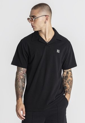 Muškarac s kratkom kosom i tetovažama na obje ruke, nosi sunčane naočale i crnu polo majicu s kratkim rukavima s malim logotipom na prsima.
