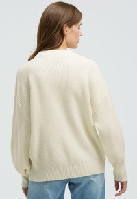 Cremfarbener, gerippter Pullover mit lockerer Passform, fallenden Schultern und langen Ärmeln. Der Stoff wirkt weich mit einer subtilen Textur. Lässiger Stil.