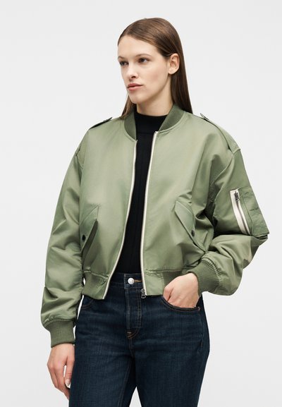 Γυναίκα που φοράει χακί μπομερ μπουφάν πάνω από μαύρη μπλούζα και σκούρα μπλε τζιν, με το ένα χέρι στην τσέπη, κοιτάζοντας προς τα πλάγια.