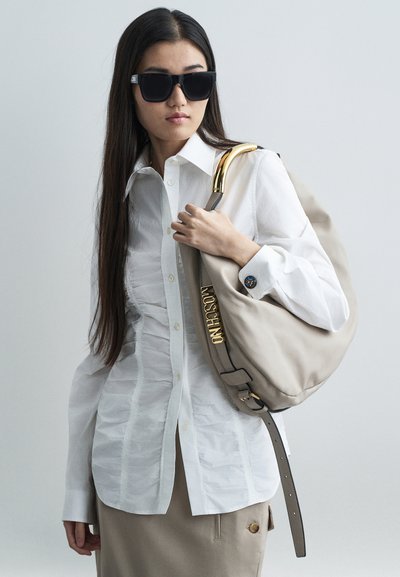 Sieviete baltā ručotajā krekls un bēša svārki, kas nēsā lielas melnas saulesbrilles, tur bēšu Moschino rokassomu ar zelta furnitūru uz pleca.