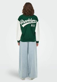 Zielono-biała kurtka varsity z napisem "Brooklyn N.Y." na plecach, w zestawie z jasnoniebieskimi szerokimi spodniami dżinsowymi. Miękki materiał, luźny krój.