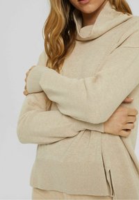 Kvinna med långt blont hår klädd i en beige stickad tröja med hög krage, med armarna korsade över bröstet framför en enfärgad bakgrund.