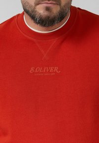 Punane dressipluus ümarate lõhikute, soonikuvarrukate ja -äärtega. Rinna peal on trükitud tekstilogole "SOLIVER" veidi tumedamas varjus.