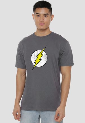 Young man wearing a gray t-shirt with a yellow lightning bolt inside a white circle, standing against a plain light background.