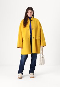Cappotto di lana giallo con grandi bottoni, vestibilità oversize, tasche laterali; abbinato a un top blu, jeans scuri e sneakers beige. Inclusa borsa bianca.