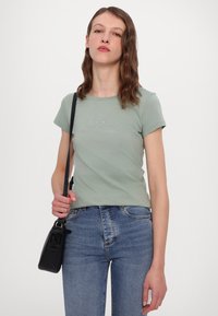 Lichtgroene t-shirt met korte mouwen, voorzien van een subtiele logo in rhinestones, gecombineerd met blauwe denim jeans en een kleine zwarte schoudertas.