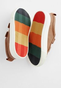Chaussures en daim brunes avec des sangles velcro ; semelles en caoutchouc blanches présentant un motif rayé multicolore en texture chevrons sur la semelle extérieure.