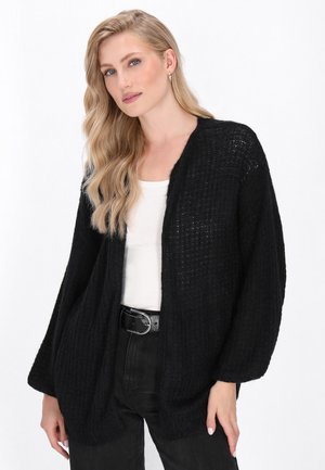 Cardigan in maglia oversize nera con una vestibilità morbida, apertura frontale e motivo testurizzato. Abbinato a una maglietta bianca e pantaloni neri.