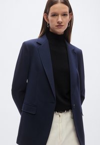 Granatowa marynarka o dopasowanym kroju, z pojedynczym zapięciem na guzik i rozciętymi klapami. Noszona na czarnym golfie, z widocznym kremowym pasem w talii.