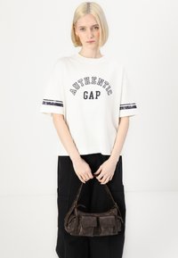 Bílý bavlněný tričko s potiskem "AUTHENTIC GAP" v námořnické modři, s krátkými rukávy a tmavými pruhy. Tmavá kabelka s kapsami je držena v rukou.
