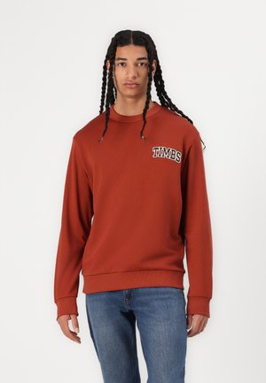 Bruine sweatshirt met lange mouwen, voorzien van geribde manchetten en zoom, met het "TIMBS" logo in het wit gedrukt op de linkerborst. Gepaard met blauwe jeans.