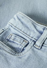 Gai zila džinsu tuvplāns, kurā redzama detalizēta šuvju apdare un mazs priekšējais monētu kabata virs galvenās kabatas.