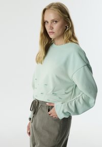 Sudadera verde menta de corte relajado, con detalles bordados y puños acanalados. Combinada con pantalones elásticos de cintura en gris claro.