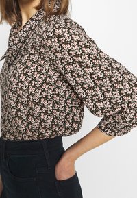 Blusa nera floreale con fiori rosa e bianchi, dotata di colletto, maniche ampie e tessuto testurizzato. Abbinata a jeans scuri.