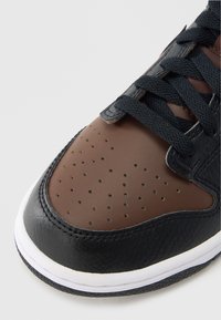 Adidaș din piele neagră cu vârf perforat maro, șireturi negre și talpă de cauciuc albă. Materialele texturate sporesc aderența și structura.