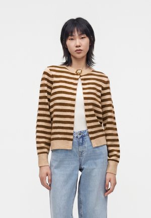 Jeune femme aux cheveux noirs porte un cardigan beige et marron rayé, un haut blanc et un jean bleu clair sur un fond blanc uni.