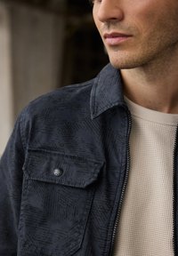 Giacca blu navy con texture, colletto e tasca frontale, con un sottile motivo geometrico e una cerniera, indossata sopra una camicia crema in maglia a nido d'ape.