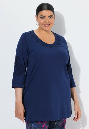 Femme aux cheveux foncés portant un haut bleu marine à manches longues avec des détails brodés, souriante devant un fond blanc uni.