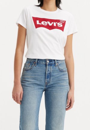 Femme portant un t-shirt blanc Levi’s et un jean bleu taille haute, debout devant un fond blanc uni.